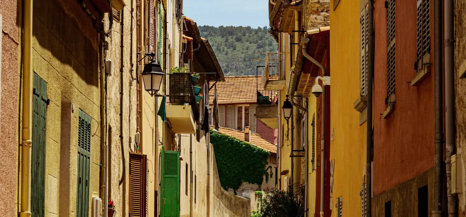 Rue en pente bordée de façades ocre et de volets verts, collines au fond